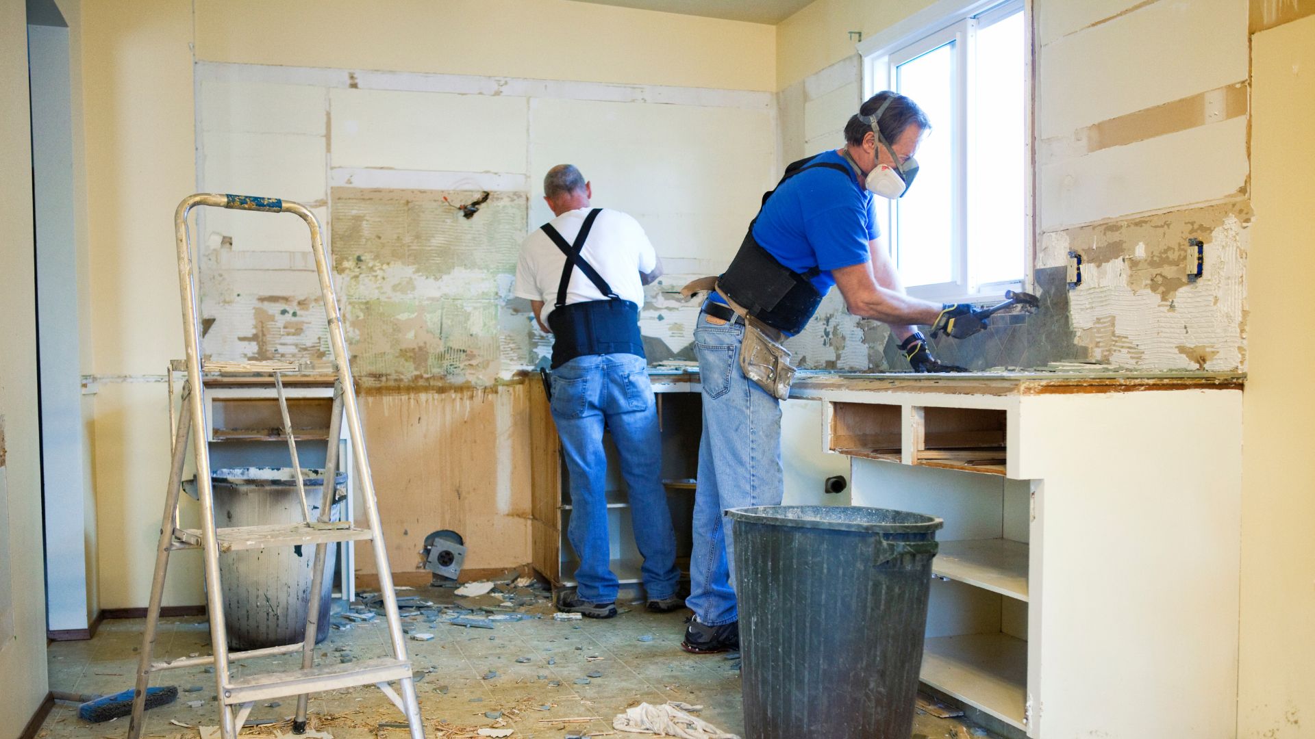 Two construction workers in protective gear renovating a kitchen, removing old cabinets and tiles.