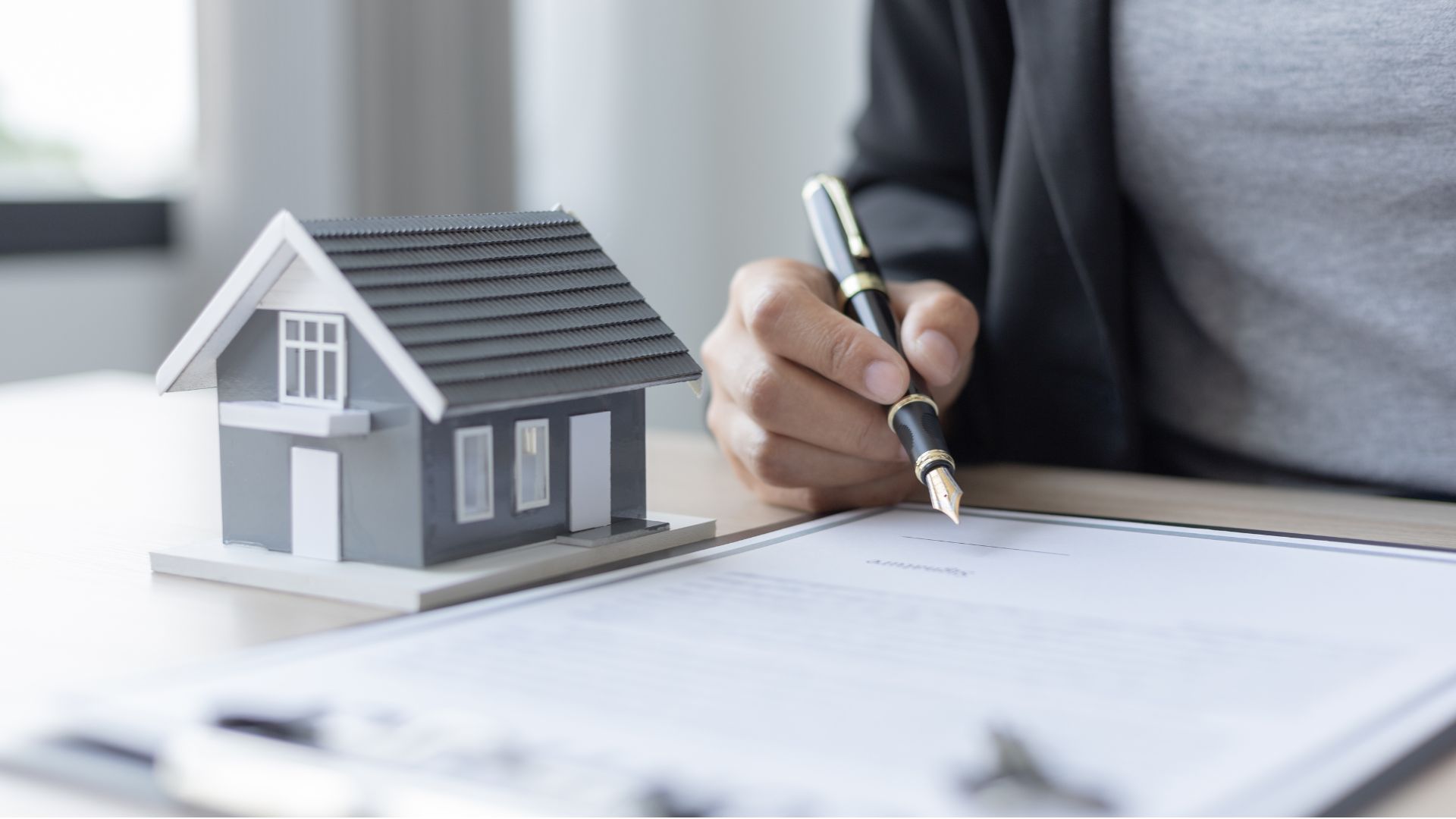 A person in a suit holding a pen, reviewing a contract with a miniature model house on the table, symbolising real estate or mortgage signing.