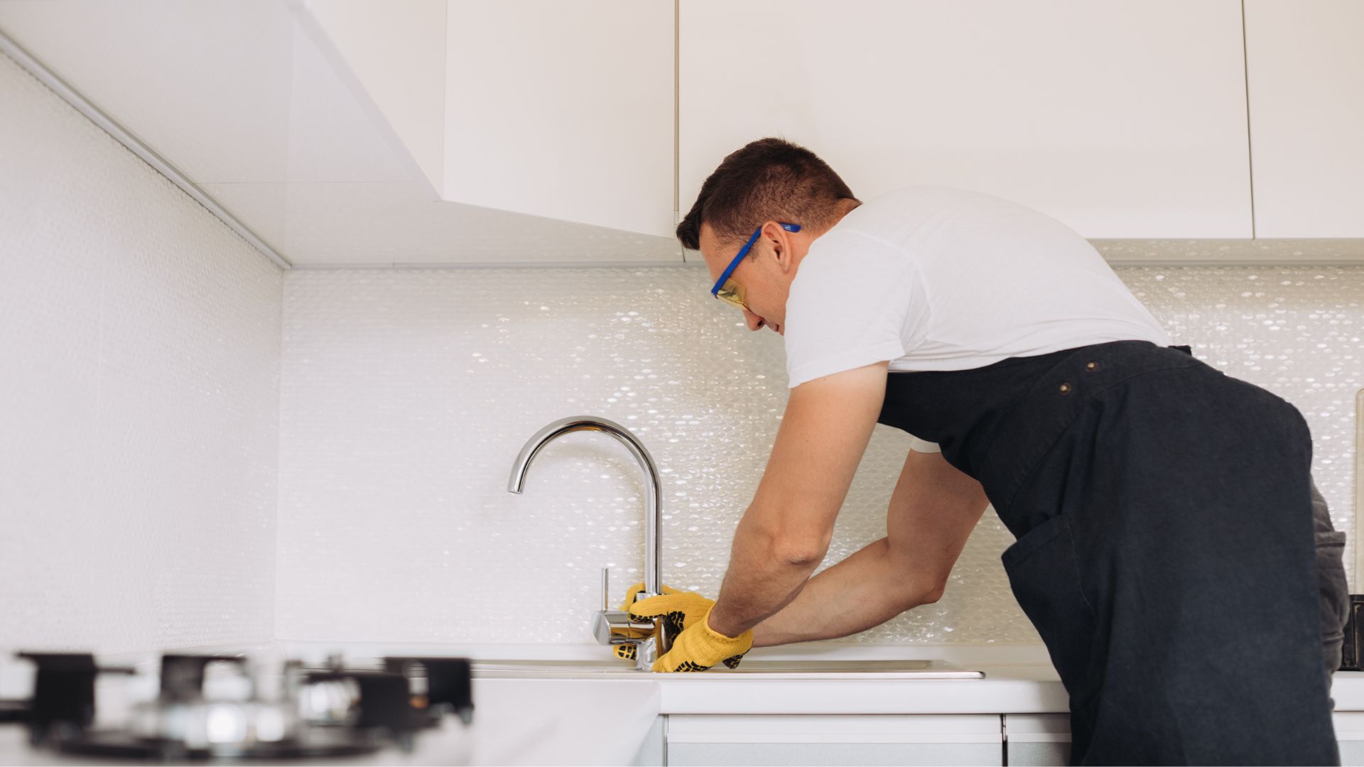 A man wearing safety glasses and gloves installing a faucet in a modern white kitchen.